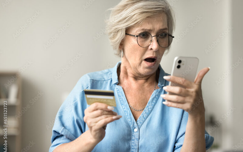 Shocked concerned mature woman in glasses holding credit card, staring at smartphone in bad surprise, getting stress, financial problems, money stealing, overspending, bankruptcy risk